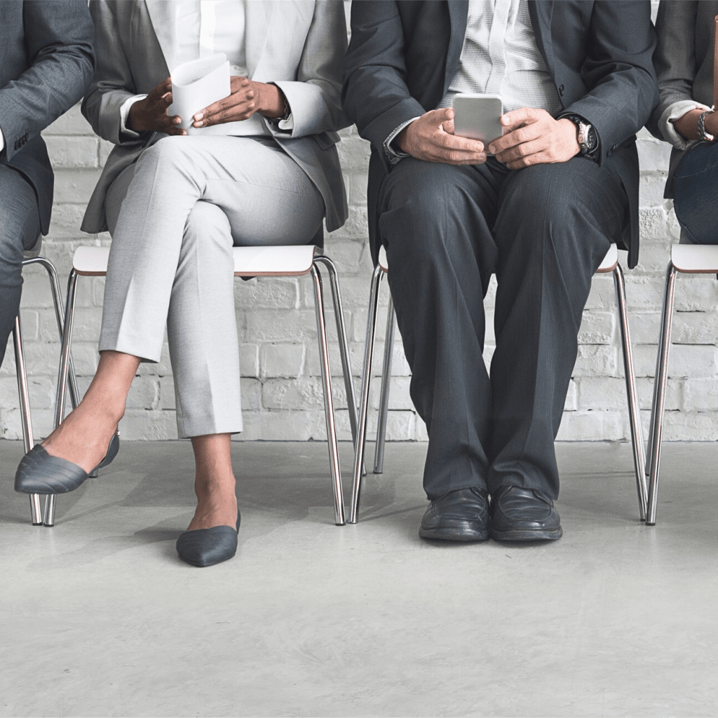 Three job candidates dressed in professional attire sit side by side in chairs, waiting for their interviews. One holds a document while another is checking a smartphone.